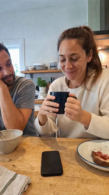 A woman smiles at her phone over a mug as her partner leans in, a real customer reaction