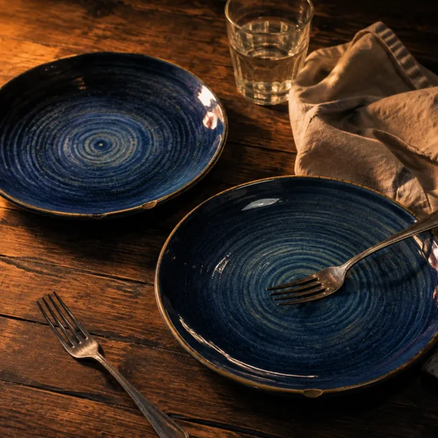 Two chipped handmade blue plates, silver forks and a linen napkin on a warmly lit wooden table