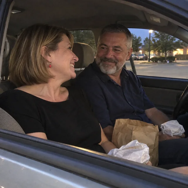 A married couple smiling at each other over takeaway burgers in a parked car; she wears small red earrings.